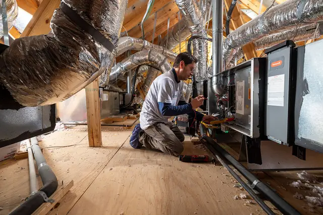 Champion and Nash technician in a branded long-sleeve shirt kneeling in a finished attic beside an air handler and a Honeywell control panel, meter leads connected