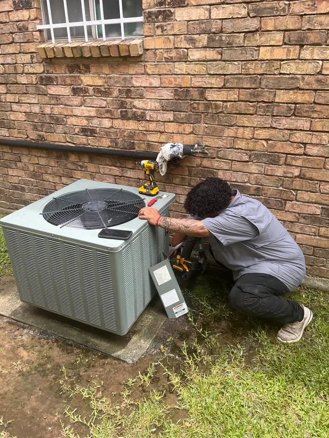 Champion and Nash technician crouched at an outdoor condenser against brick, gauges in play, driver resting on the unit
