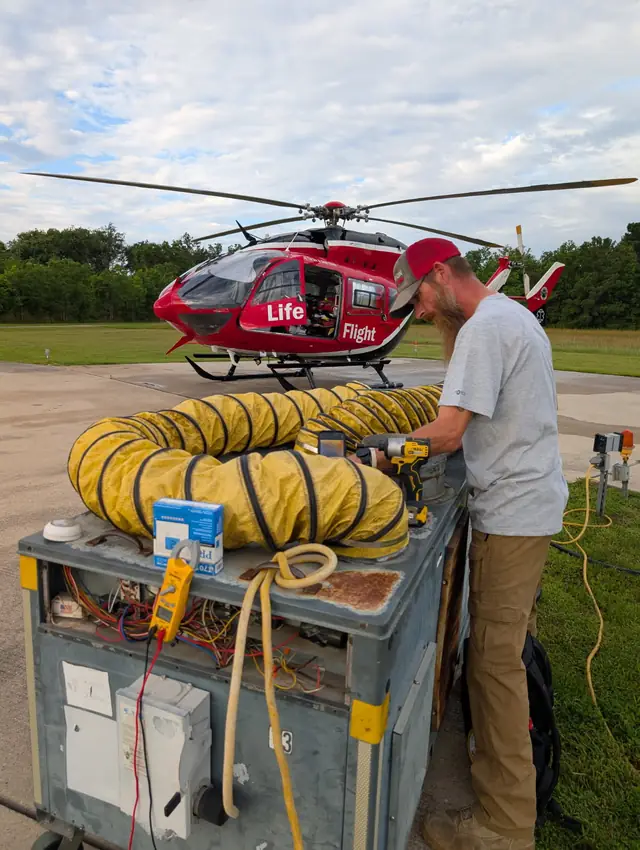 A Champion and Nash technician sets up a portable cooling unit with yellow flexible ducting run out onto a hospital helipad, an emergency medical helicopter parked behind