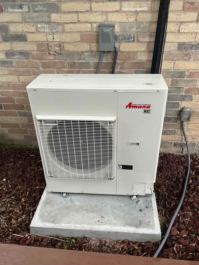 A newly installed white Amana R32 outdoor heat pump unit standing on a fresh concrete pad against a brick wall